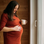 Babybauchshooting zu Hause. Hochschwangere steht am Fenster, hält Tasse in der Hand und schaut auf Babybauch den sie mit anderer Hand festhält.
