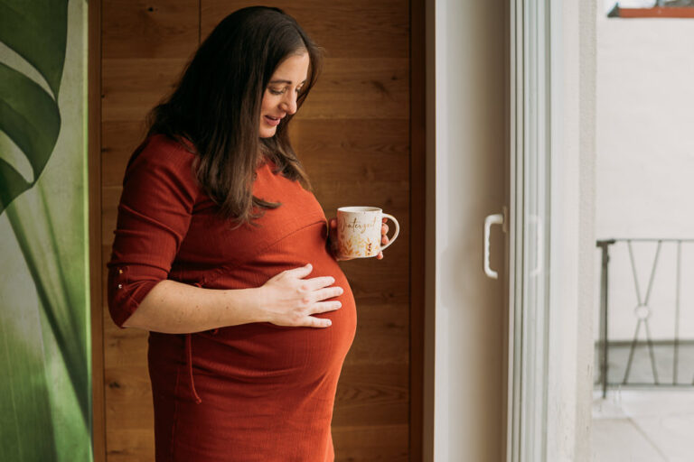 Babybauchshooting zu Hause. Hochschwangere steht am Fenster, hält Tasse in der Hand und schaut auf Babybauch den sie mit anderer Hand festhält.