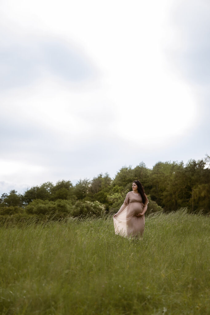 Schwangere Frau hält Babybauch fest, schaut in die Ferne und läuft über Wiese auf Fotograf zu